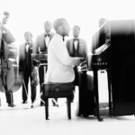 John Legend gra na fortepianie w otoczeniu muzyków w czarnych garniturach – album Muse 2026